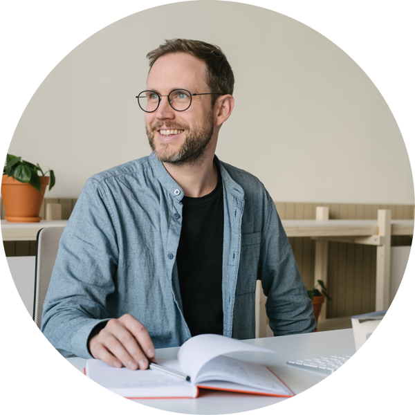 Person männlich gelesen, weiß, Brille, lächelnd, mit Notizbuch