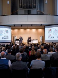 großer Saal mit Publikum und Bühne mit zwei Personen