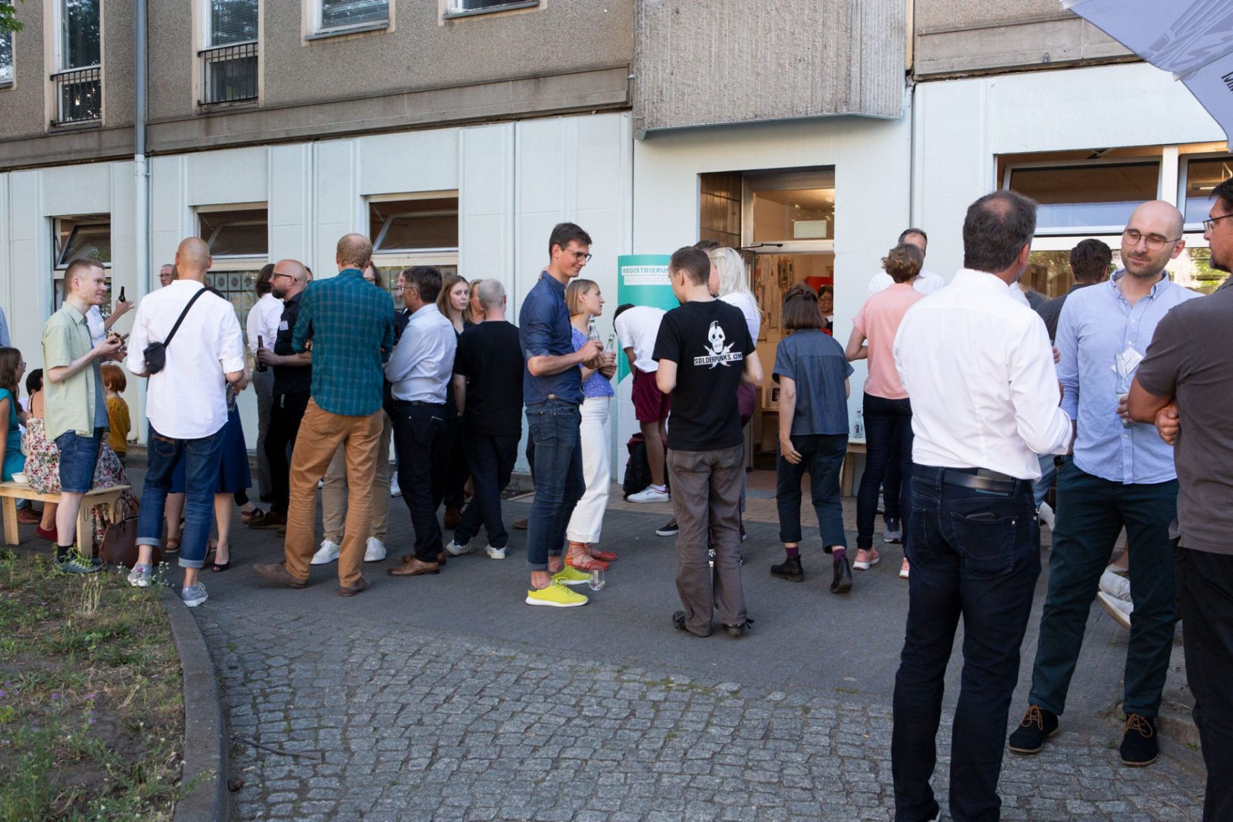 circa 20 Personen stehen vor Gebäude und unterhalten sich