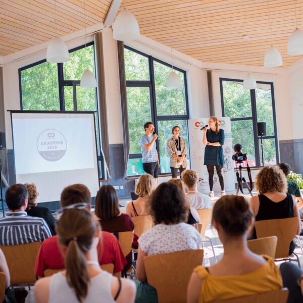 Event C2C Academy, three people on bright stage, audience sitting in the foreground