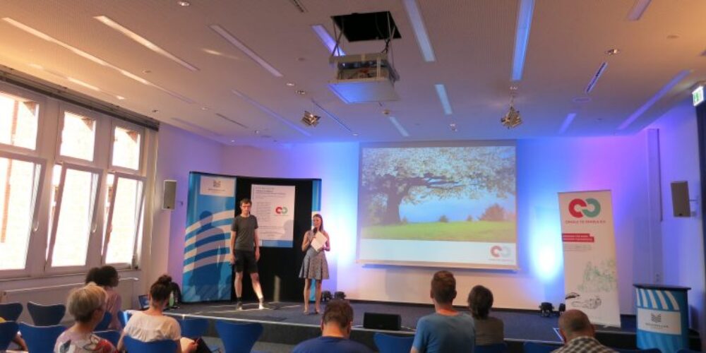 Saal mit Publikum und zwei Personen auf Bühne mit Präsentation