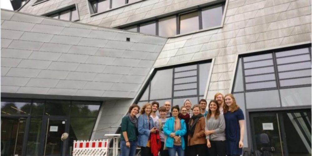 Gruppe von ca. 15 Menschen vor großem grauen Gebäude mit schiefen Fenstern